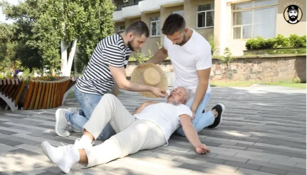Two men assist senior man on ground outside building.