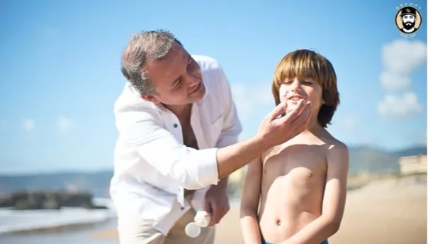 A smiling dad applies sunscreen to his son's face on a sunny beach day.