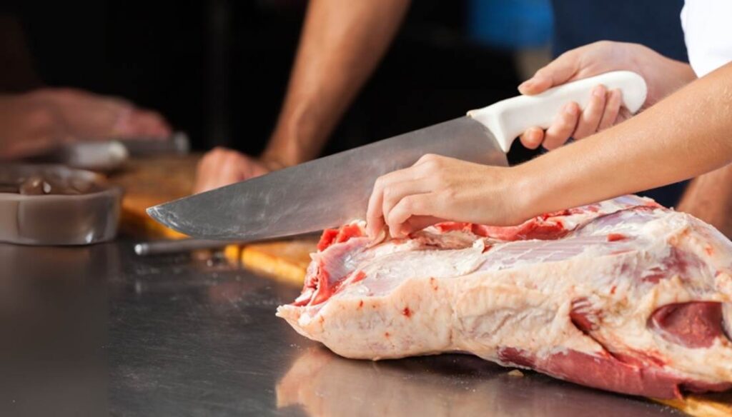 A person cutting raw meat with a large butcher knife on a metal surface.

