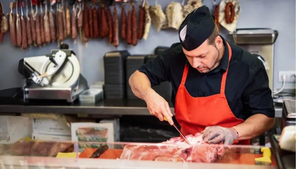 Butcher wearing a black shirt, red apron, and protective glove carefully cutting meat at a butcher shop counter, with hanging sausages and cured meats in the background.