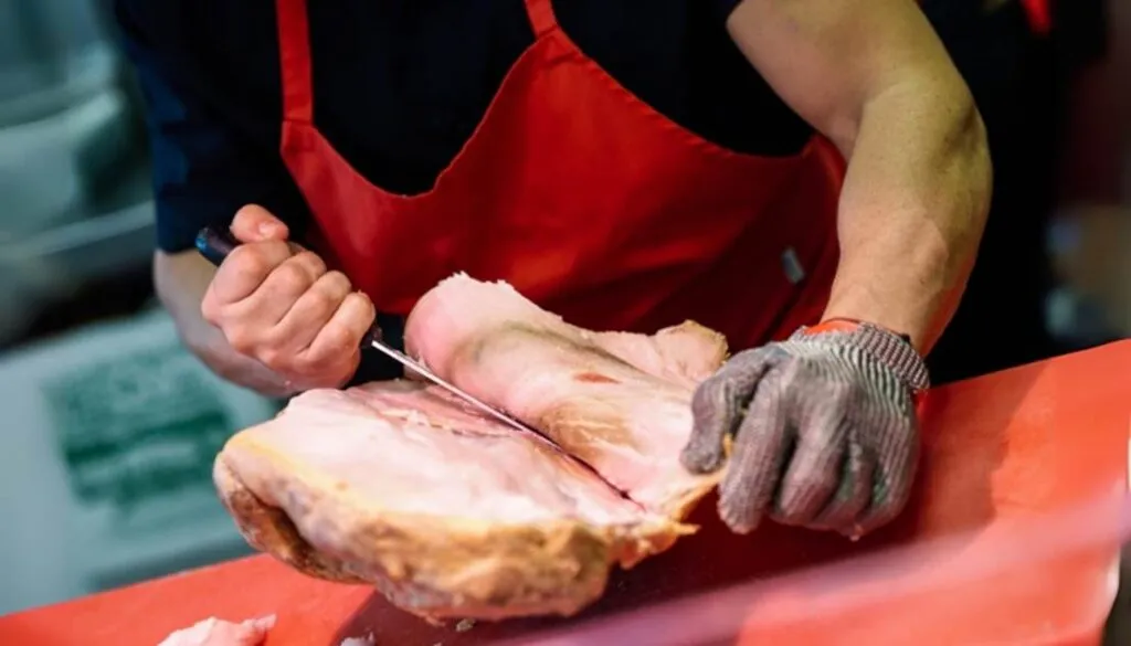 Butcher slicing a large piece of cured meat, wearing a red apron and safety gloves.
