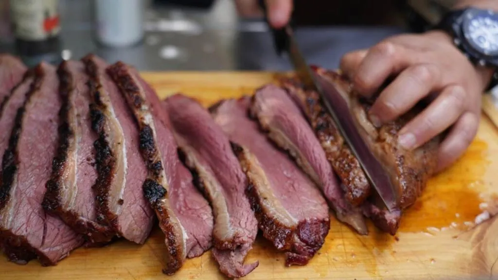 A person slicing smoked brisket on a wooden cutting board, revealing juicy, tender slices with a dark, seasoned bark on the edges.