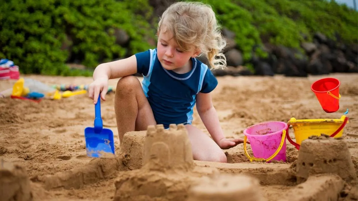 Child building sandcastle on beach with colorful buckets.